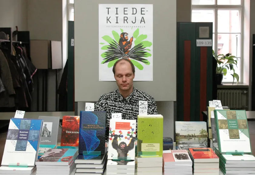 Pöydällä on esillä kirjoja. Pöydän takana istuu mies, ja hänen takanaan on juliste jossa lukee Tiedekirja.