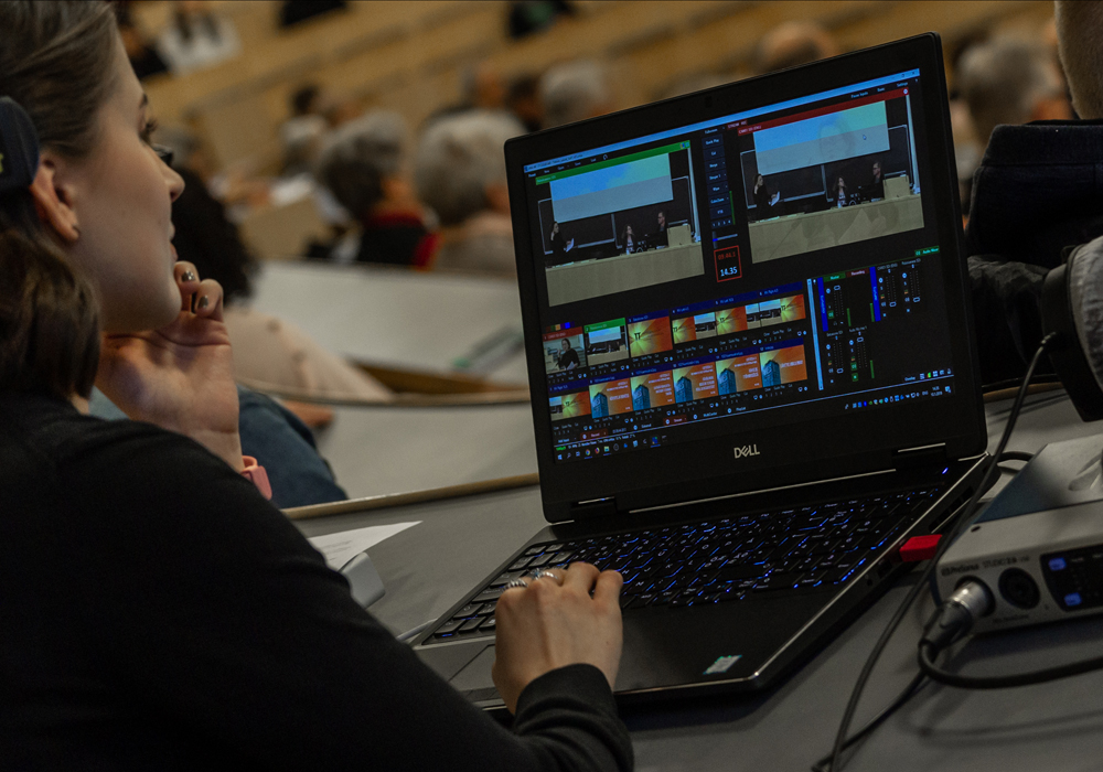 Närbild på person vid bärbar dator på vars skärm hen följer föreläsningens lagring på video. I bakgrunden syns publik i föreläsningssalen.
