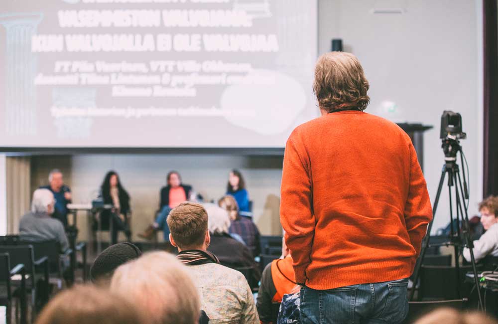 Punaiseen paitaan pukeutunut henkilö on noussut seisomaan luentosalissa pitääkseen puheenvuoron. Hänen edessään lavalla näkyy panelisteja istumassa.