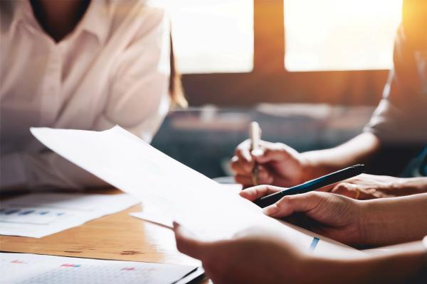 Kuvassa ihmisten käsiä, jotka pitelevät muistiota ja kynää, taustalla näkyy läppäri.