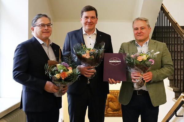 Kolme miesoletettua henkilöä puvut päällä ja kukkakimput käsissään.